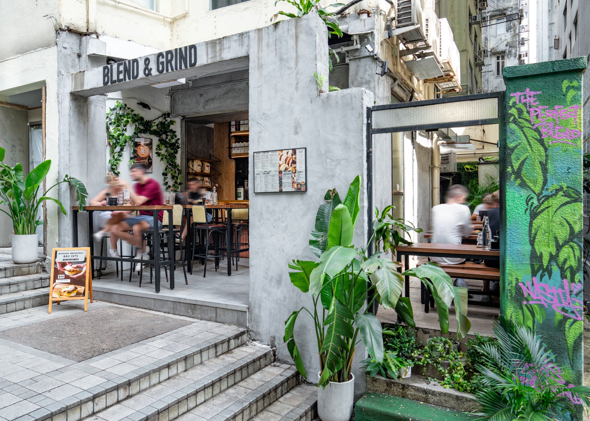 Blend & Grind’s Mid-Levels storefront beside steps, with outdoor seats, tropical plants and a painted green wall.