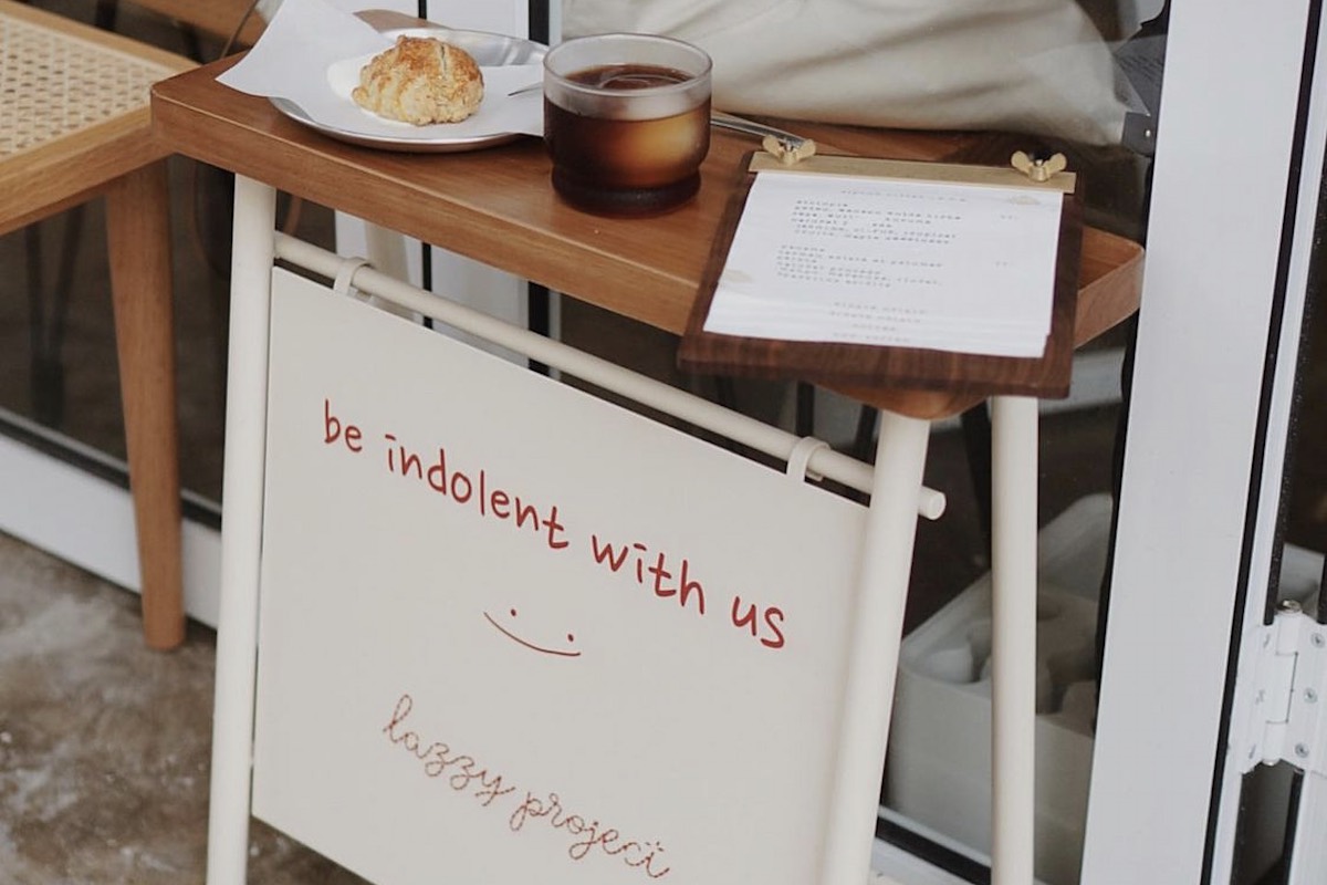 Lazzy Project’s entrance table with coffee and a scone above its handwritten welcome sign, beside wooden seating.