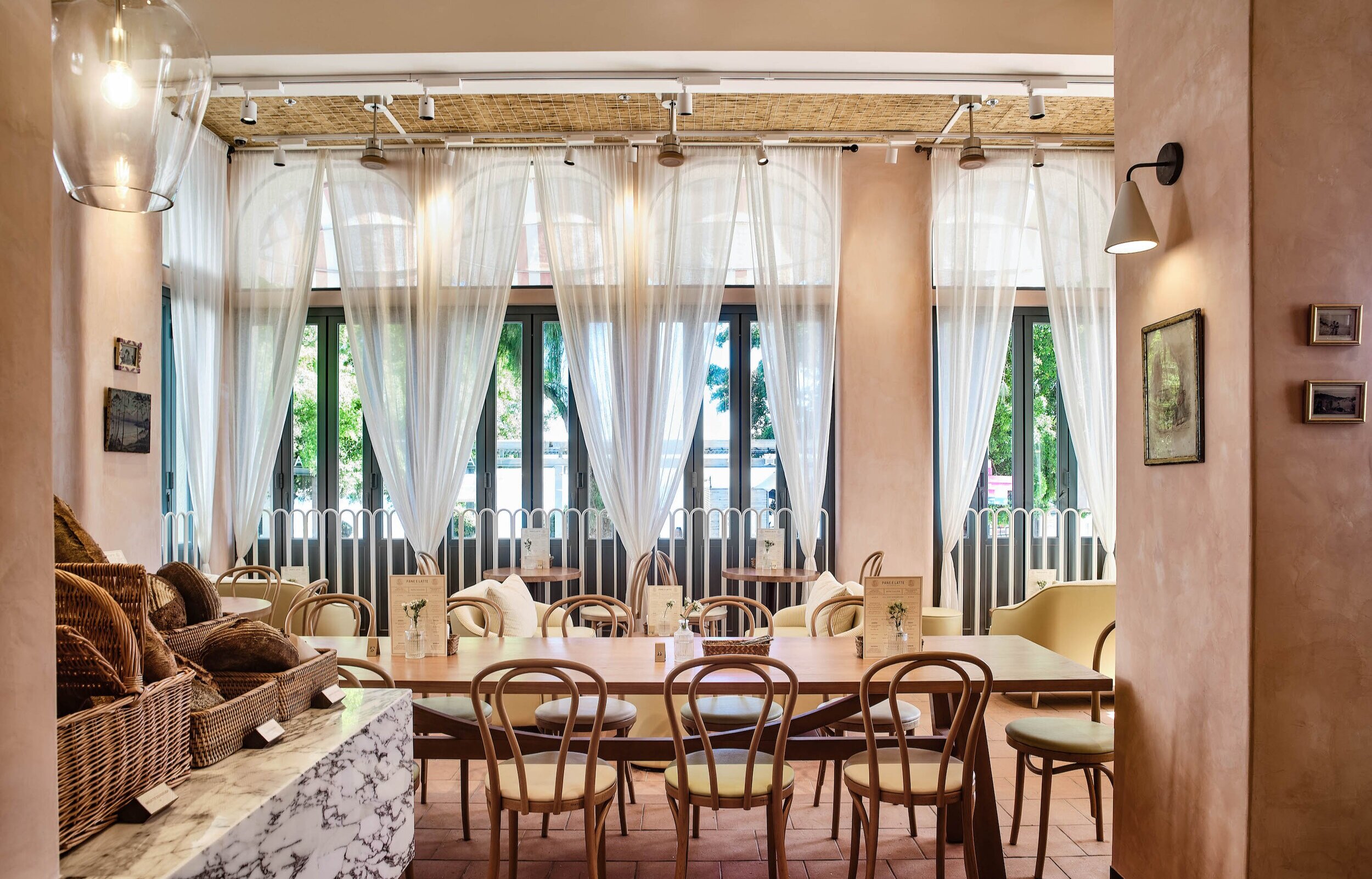Pane e Latte’s Stanley dining room with pink columns, tall white curtains, wooden chairs and bakery baskets.