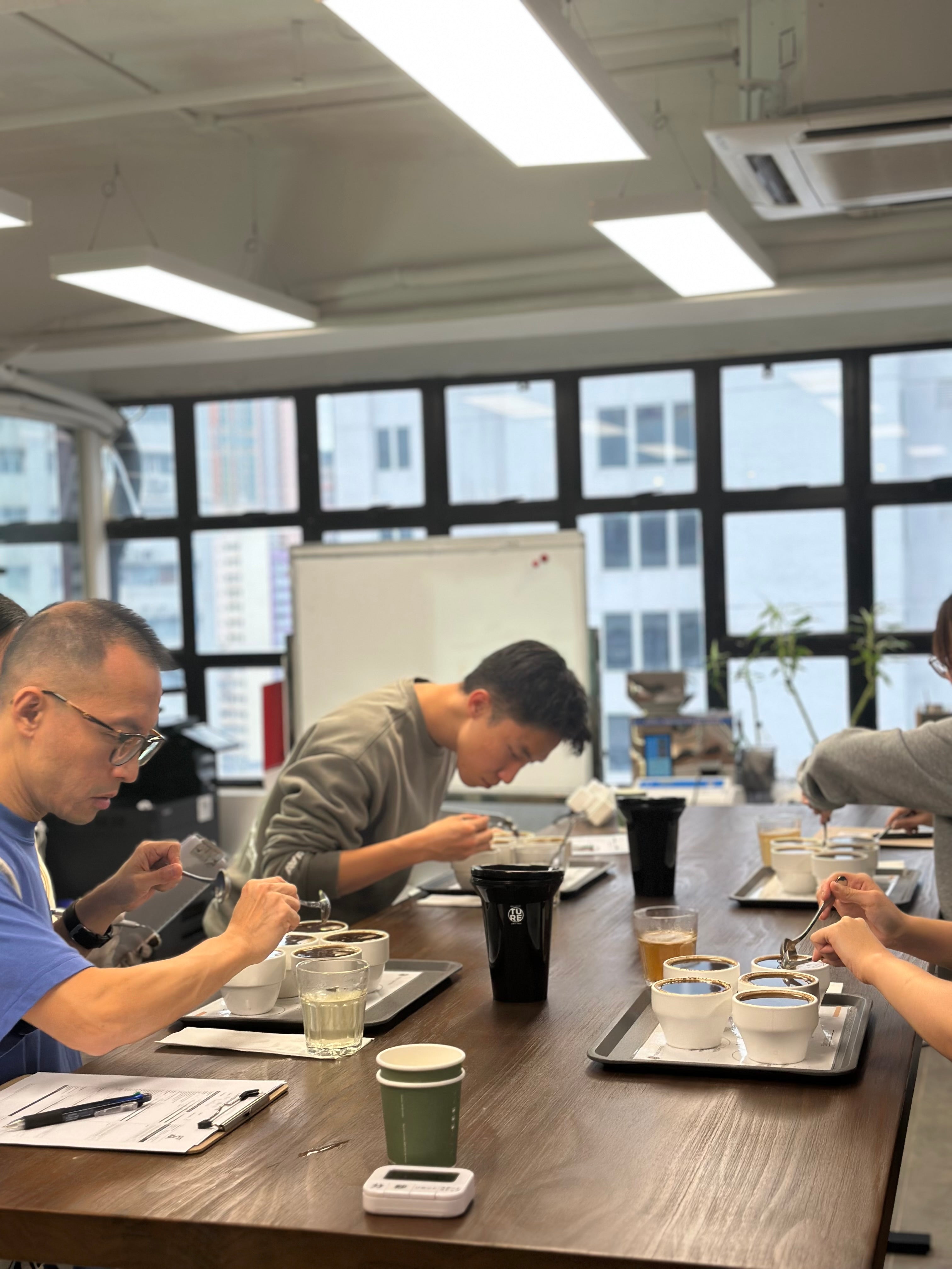 A coffee cupping class at HOLISTIC’s Kwai Chung roastery, with tasting bowls on a long table beside industrial windows.